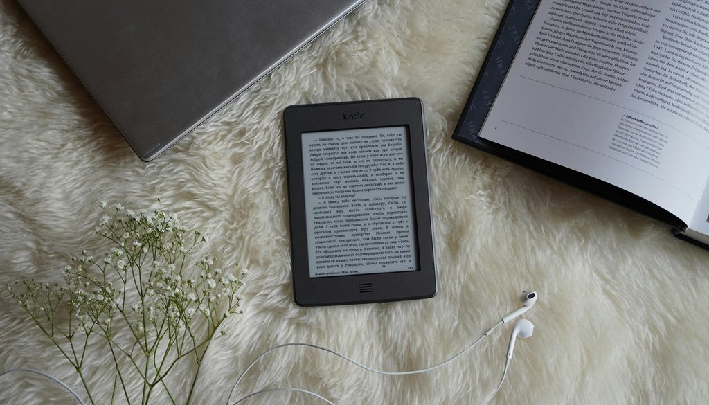 Leitor e-reader sobre um tapete felpudo, ao lado de livro impresso, revista e fones de ouvido, representando diferentes formatos de leitura digital e impressa