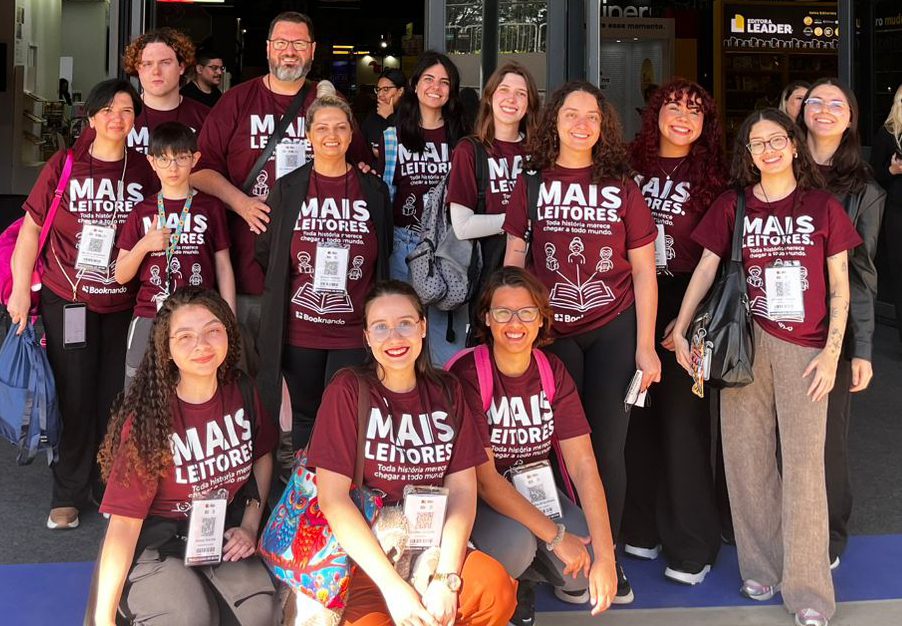 Quinze integrantes da equipe Booknando reunidos em uma feira do livro, com camisetas bordô estampadas com a frase Mais leitores: toda história merece chegar a todo mundo.
