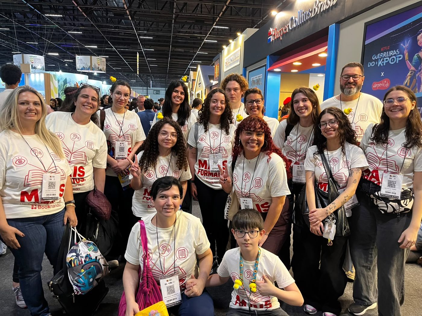 Quinze integrantes da equipe Booknando reunidos no corredor de uma feira do livro, com camisetas brancas estampadas com a frase “Mais formatos: um livro, muitas formas de ler”.